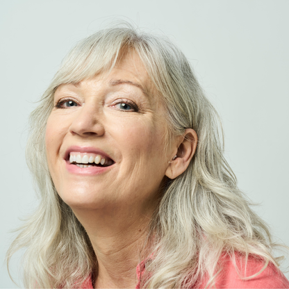 A smiling older woman with long, wavy gray hair and light skin wears a pink top against a plain background, discreetly enjoying Ceretone Core One Pro Invisible-In-Canal Hearing Aids.