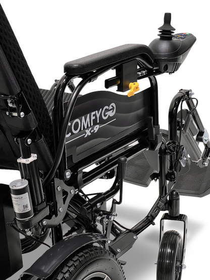 Close-up of a black ComfyGo X-9 Remote Controlled Powerchair - Automatic Reclining, featuring the joystick on the armrest. Its detailed mechanical frame and rear wheel highlight precision engineering in this innovative mobility solution.