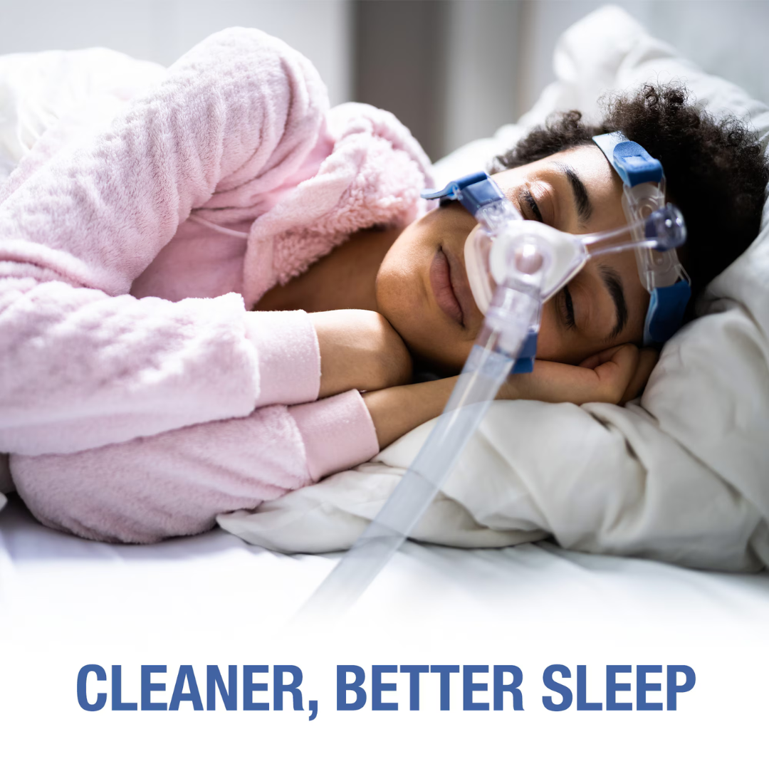 A person in a pink robe sleeps on a bed using a CPAP mask connected to a hose. The text reads: CLEANER, BETTER SLEEP—thanks to regular CPAP equipment cleaning with DMI CPAP Mask Wipes - XL Hypoallergenic & Unscented.