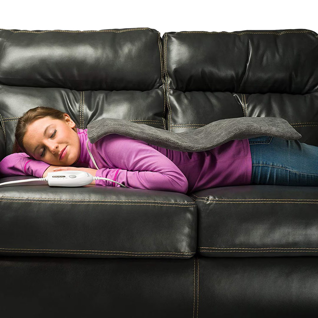 A woman lies face down on a black leather couch, eyes closed, using the DMI Dry and Moist Heat Electric Heating Pad on her back, holding its control in her hand. She is dressed in a purple sweater and jeans.