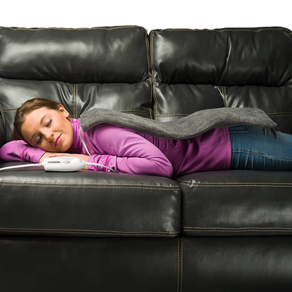 A woman lies face down on a black leather couch, eyes closed, using the DMI Dry and Moist Heat Electric Heating Pad on her back, holding its control in her hand. She is dressed in a purple sweater and jeans.