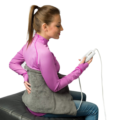 A woman in a pink top sits on a black bench, holding a controller and using the DMI Dry and Moist Heat Electric Heating Pad wrapped around her lower back for pain relief.