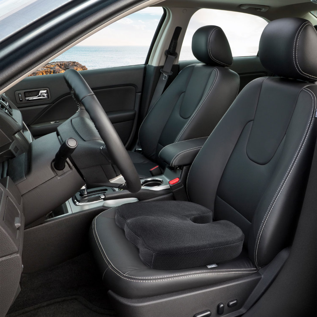The DMI Premium Coccyx Seat Cushion, crafted from black memory foam, sits on the driver’s seat and matches the black leather interior, while an ocean view is visible through the car window.