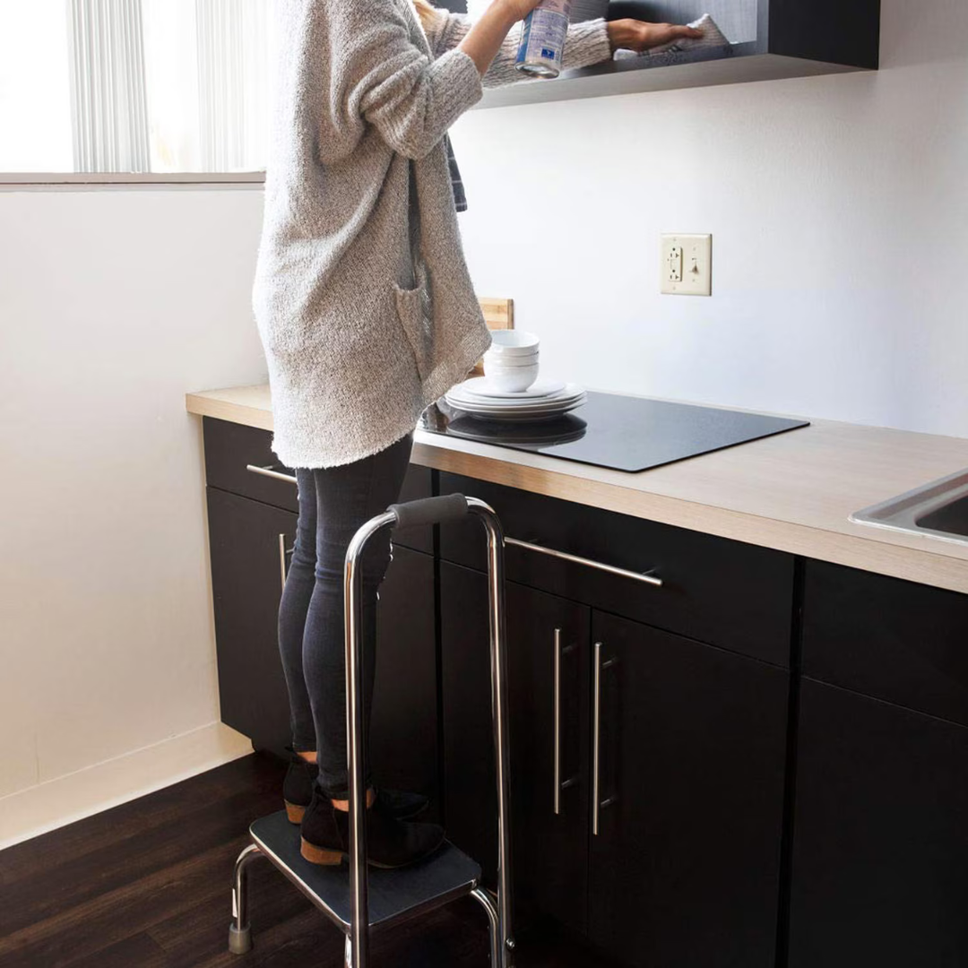 In a kitchen, someone uses the DMI Step Stool and Non Skid Rubber Platform with handrail to clean an overhead cabinet. Their face is hidden, and plates are stacked on the nearby counter.