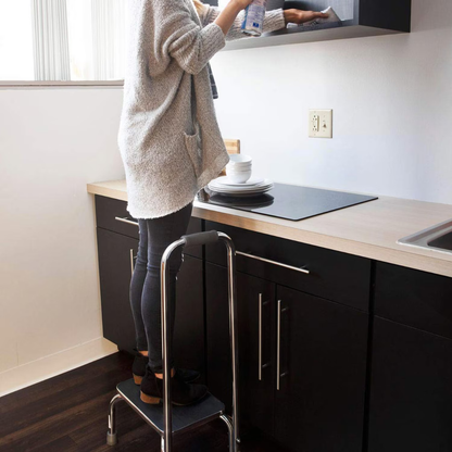 In a kitchen, someone uses the DMI Step Stool and Non Skid Rubber Platform with handrail to clean an overhead cabinet. Their face is hidden, and plates are stacked on the nearby counter.