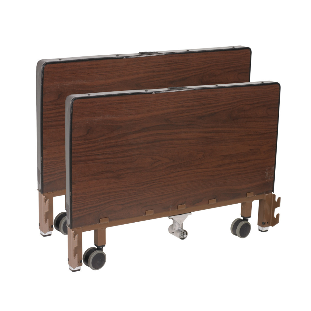 Two rectangular, wood-grain hospital overbed tables with caster wheels and metal/plastic frames are stacked vertically for storage beside a Drive Delta® Ultra-Light 1000 Bariatric Full-Electric Low Bed.