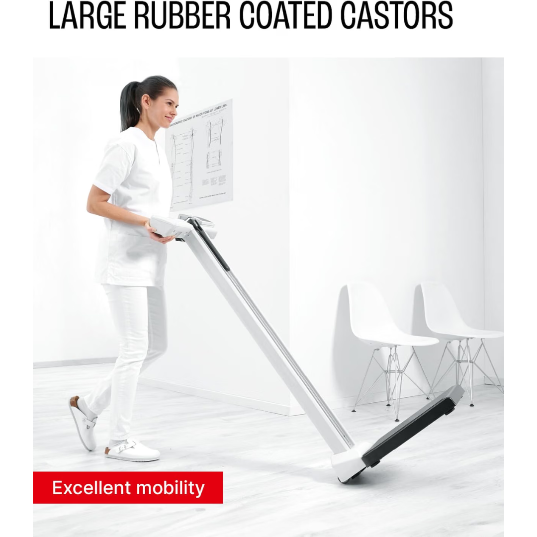 A woman in white pushes a Digital Physician Scale with Height Rod | 550 lb Capacity, featuring large rubber-coated castors, across a white room furnished with chairs and medical diagrams.