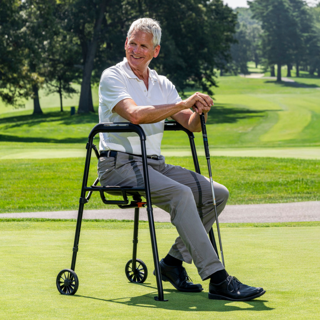 An older man sits on the Drive Air-Go Walker with Airgonomic™ Hand Comfort & Seat on a golf course, smiling as he holds a golf club. He wears a white polo shirt and gray pants, with trees and green fairways in the background.