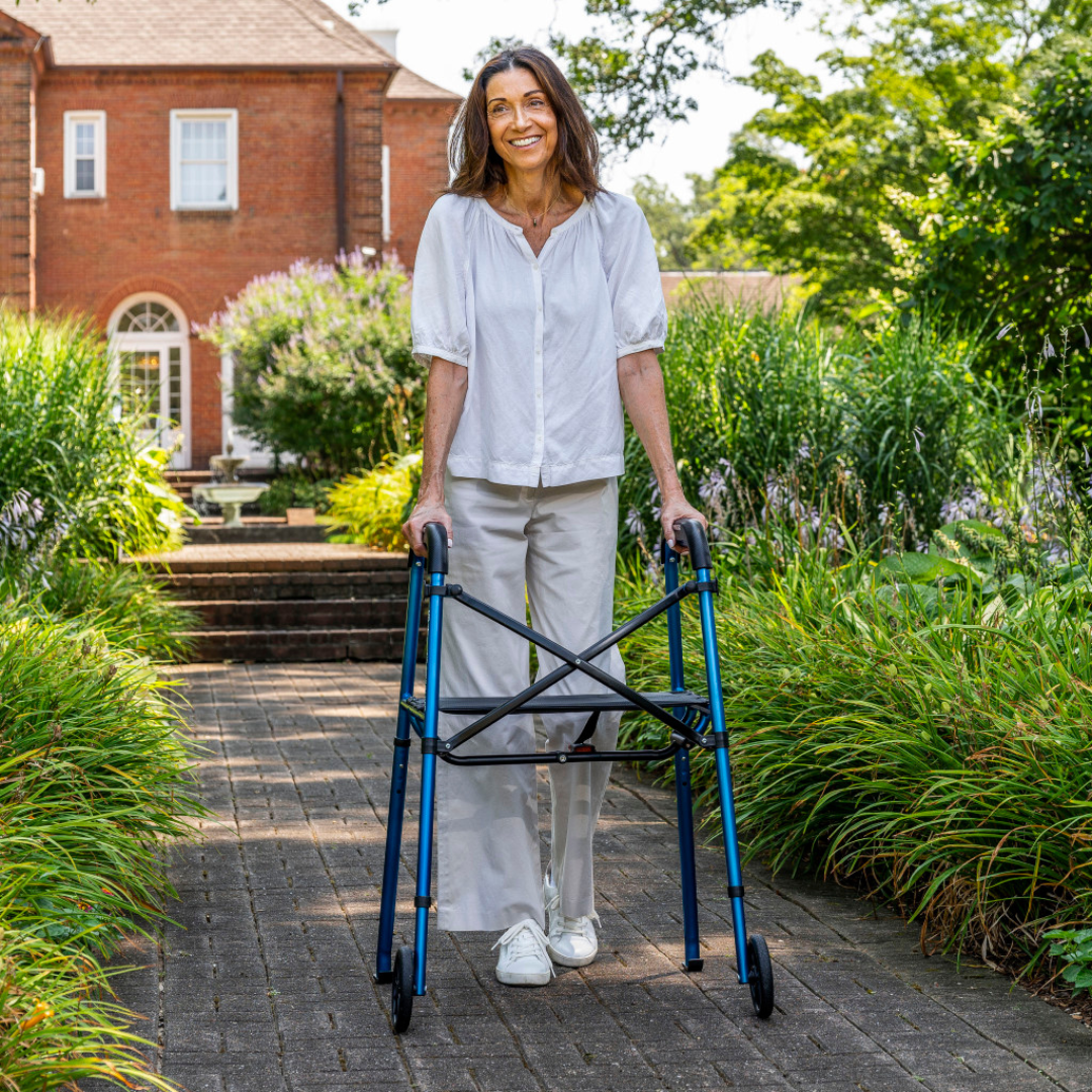 A smiling woman with long brown hair walks on a brick path in a lush garden, using the Drive Air-Go Walker with Airgonomic™ Hand Comfort & Seat, while a large brick house stands in the background on a sunny day.