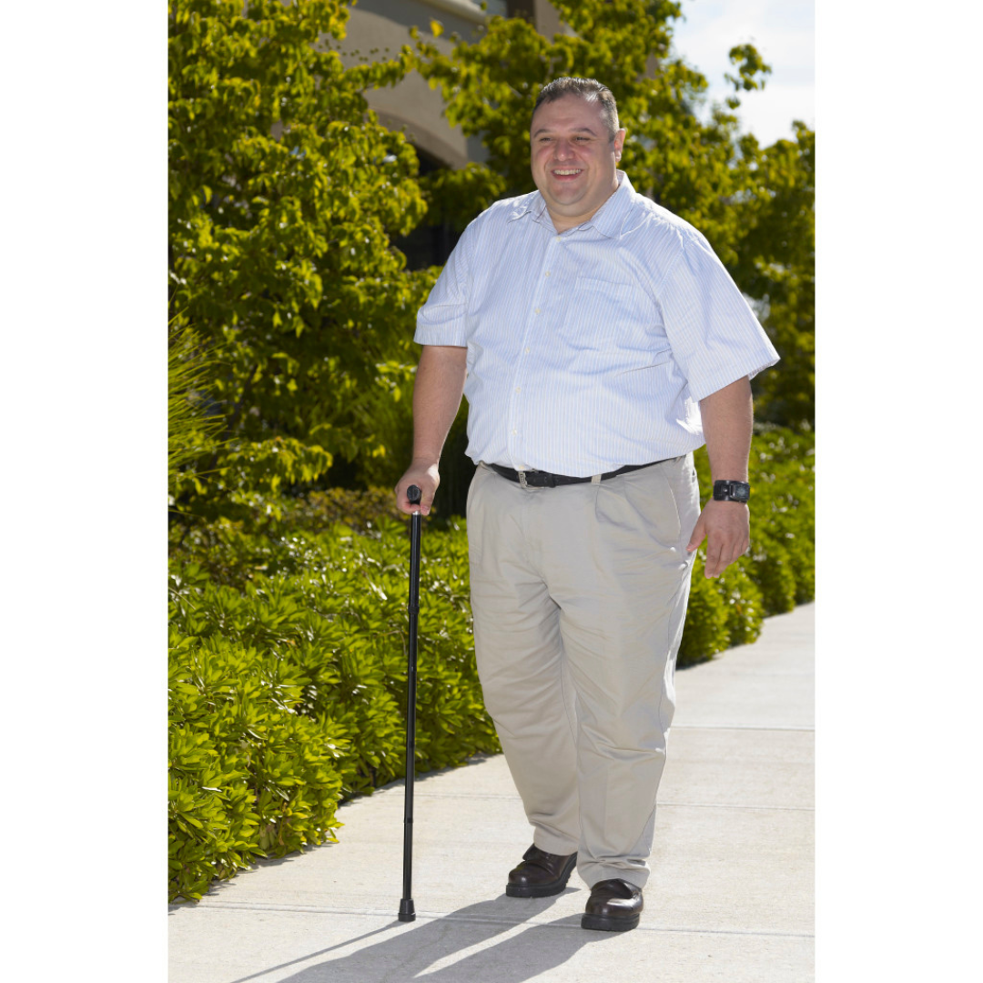 A man in light pants and a short-sleeve shirt walks on a sidewalk, smiling as he uses the Drive Bariatric Aluminum Folding Walking Cane for support. Greenery and trees are visible in the background.