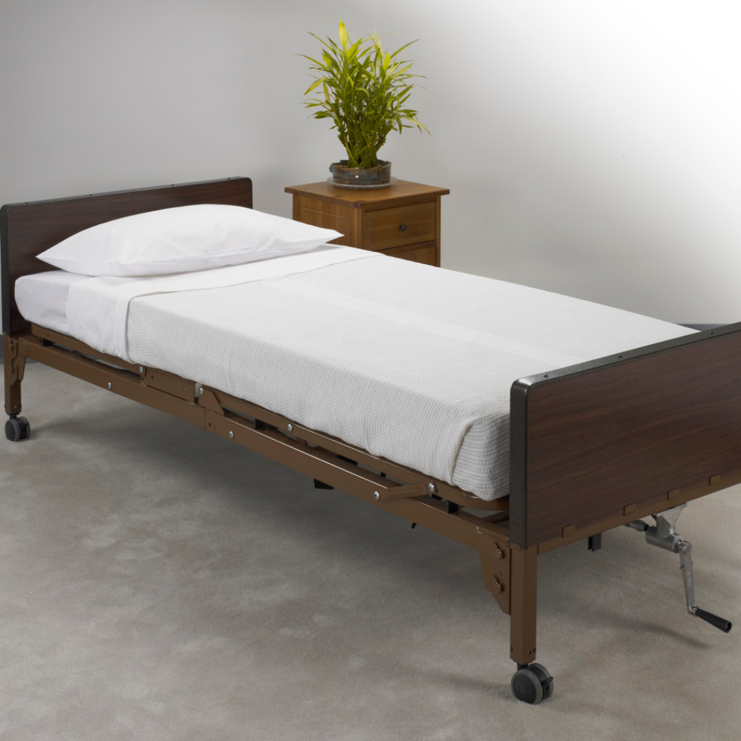 A hospital bed with a white Drive Bedding in a Box All-In-One Bedding Set and pillow is on a beige carpet. A wooden nightstand with a potted green plant is beside the bed against a plain, light-colored wall.