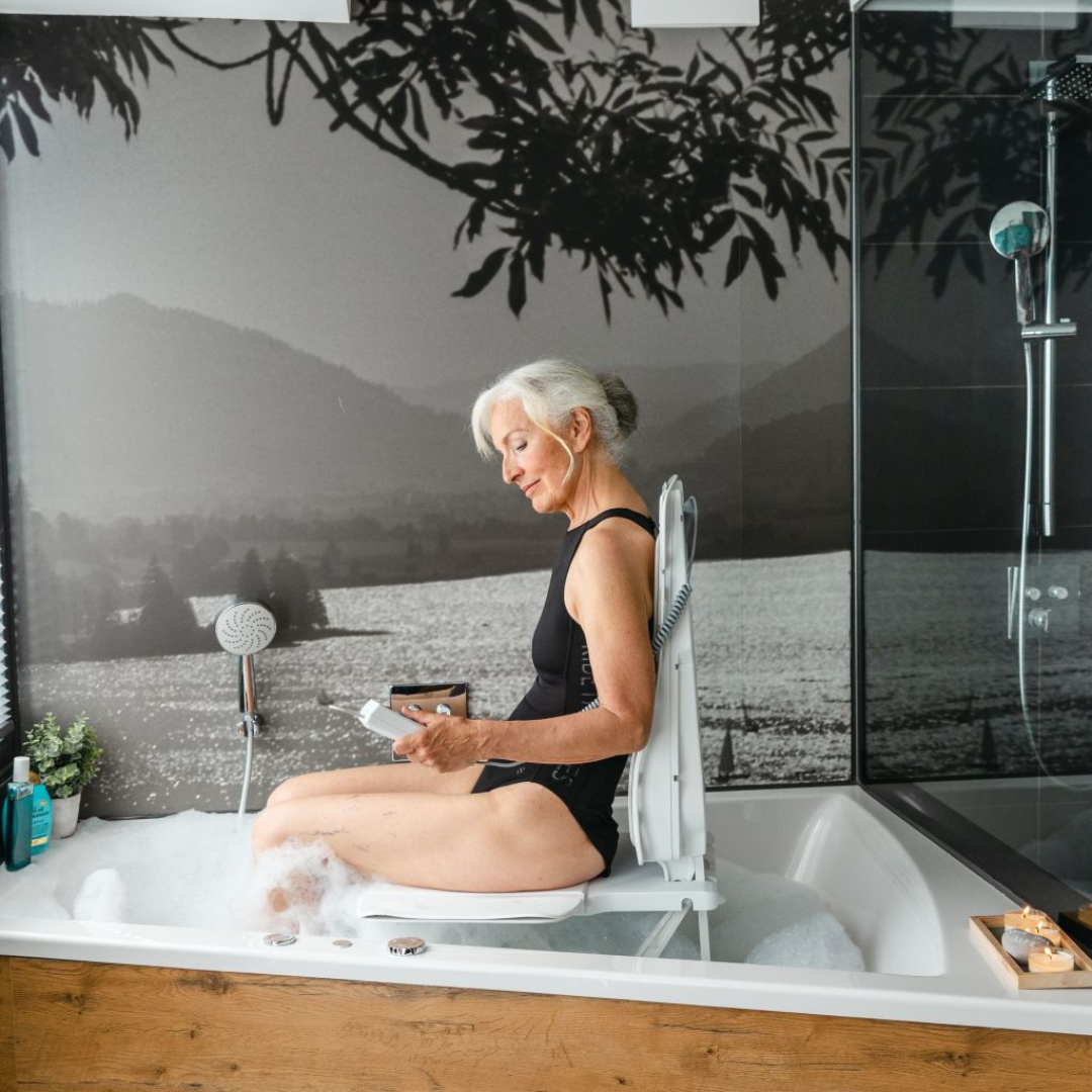 An older woman in a black swimsuit relaxes on the Drive Bellavita Dive Bath Lift—an electric reclining bathtub lift—in a bubble-filled tub, reading a book. The bathroom features a mural of a lake and mountains.