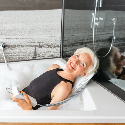 An older woman with short white hair relaxes and smiles in a bubble-filled tub, using the handheld remote of the Drive Bellavita Dive Bath Lift—an electric reclining bathtub lift—to adjust her position.