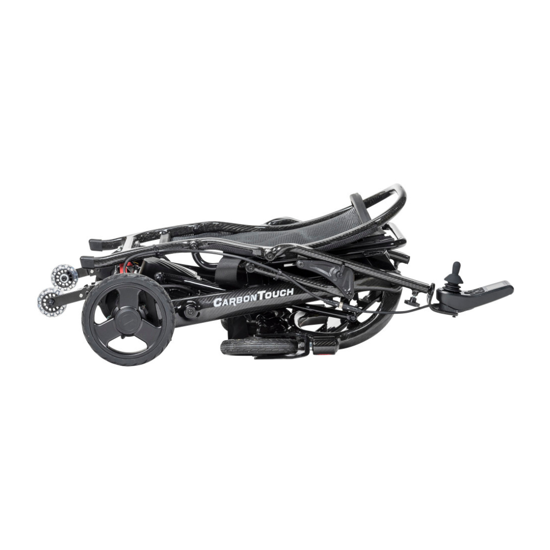 A lightweight power wheelchair, the Drive CarbonTouch Power Wheelchair is shown folded flat for transport or storage, labeled CarbonTouch. The wheels, footrest, and frame are all visible in the compact position.