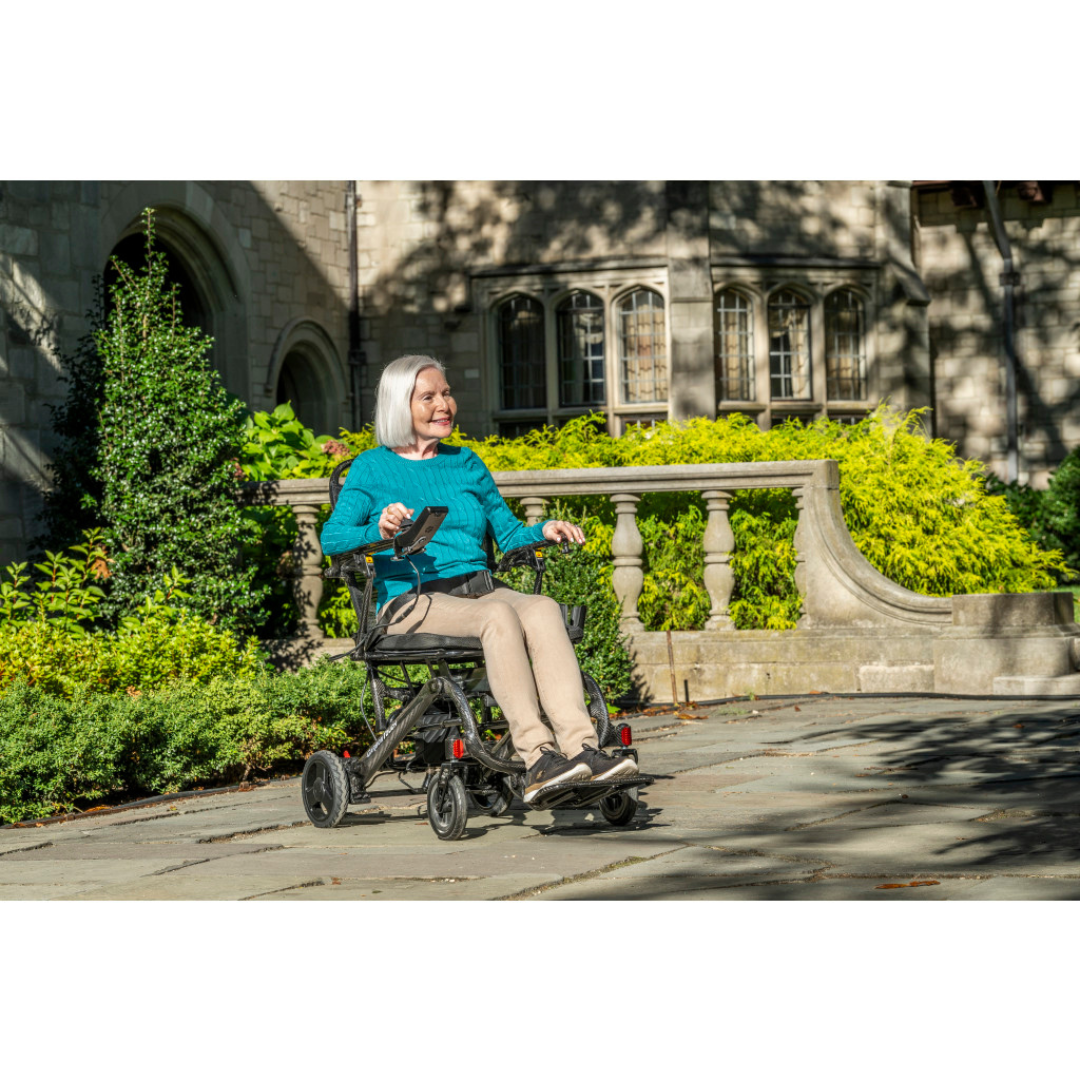 An older woman with white hair sits in a Drive CarbonTouch Power Wheelchair, smiling as she moves along a stone path outside a large building, surrounded by greenery and sunlight.