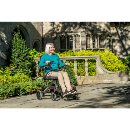 An older woman with white hair sits in a Drive CarbonTouch Power Wheelchair, smiling as she moves along a stone path outside a large building, surrounded by greenery and sunlight.