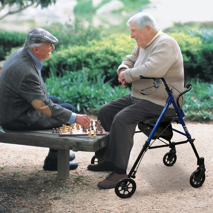 Two elderly men play chess outdoors on a bench, surrounded by greenery. One wears a cap and glasses; the other sits comfortably on his Drive Hugo Fit 6 Portable Lightweight Rollator. Both are smiling and engaged in the game.