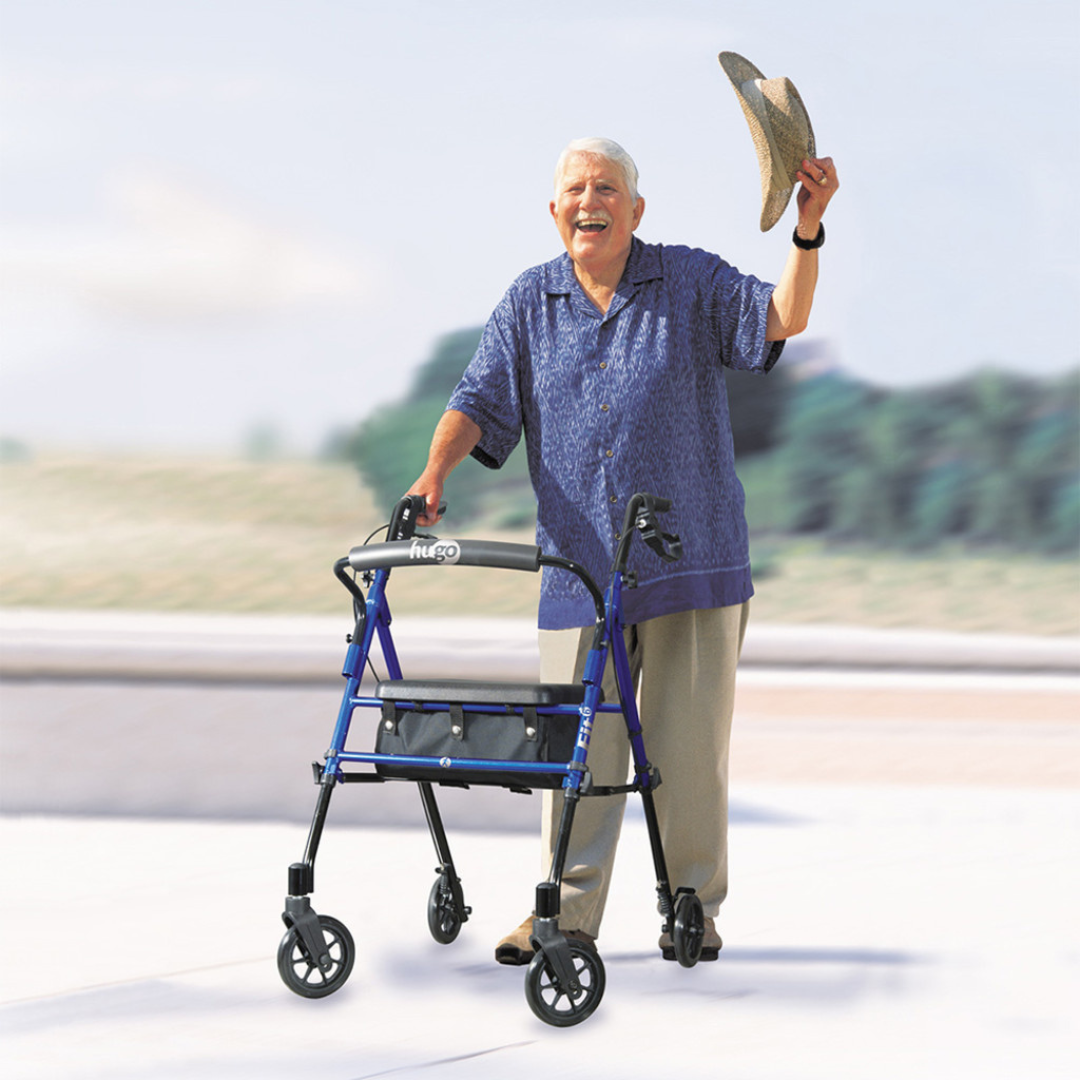 An older adult with gray hair, in a blue shirt and beige pants, smiles outdoors while using the Drive Hugo Fit 6 Portable Lightweight Rollator and waves a straw hat in the air.