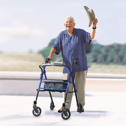 An older adult with gray hair, in a blue shirt and beige pants, smiles outdoors while using the Drive Hugo Fit 6 Portable Lightweight Rollator and waves a straw hat in the air.