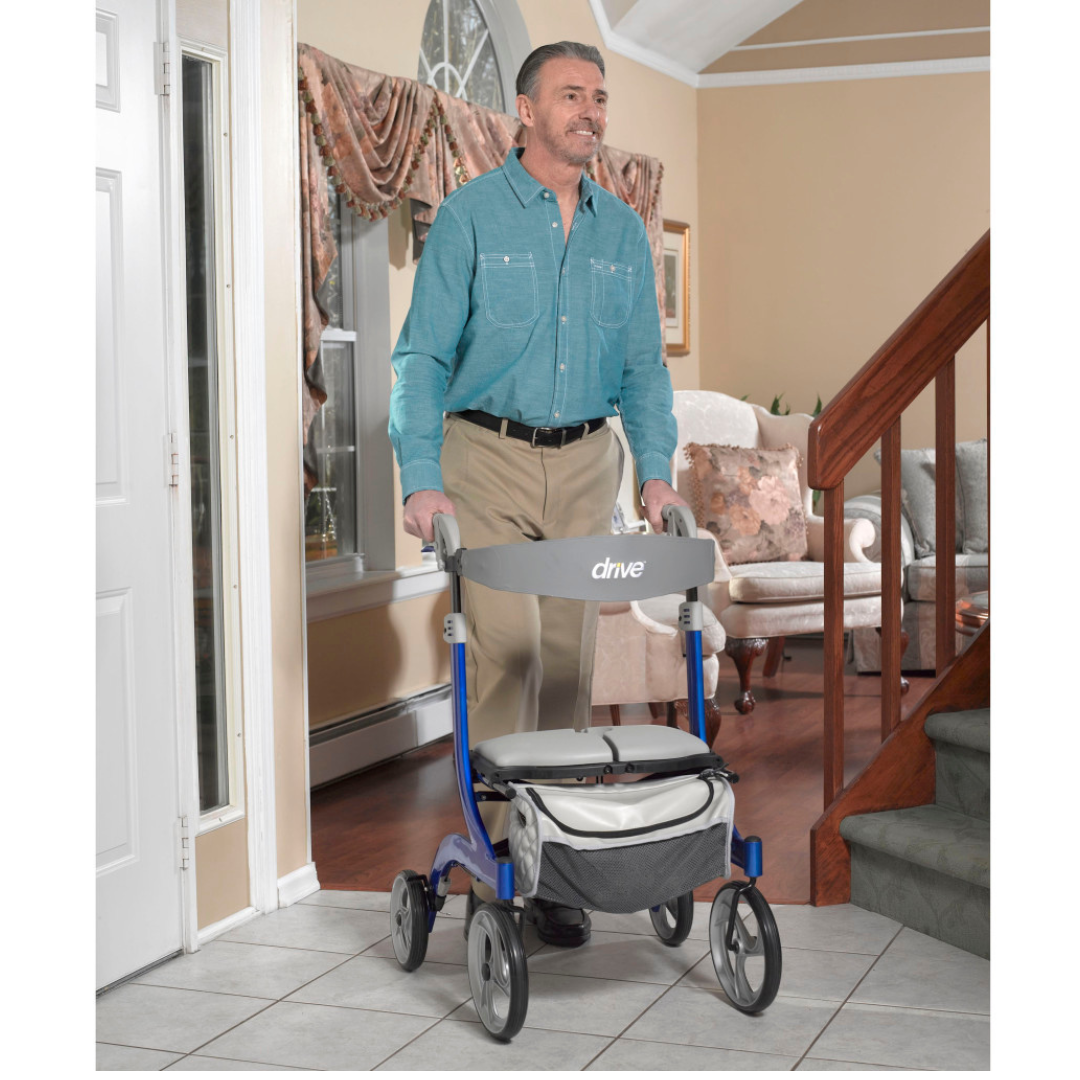 A man in a teal button-up and khakis uses the Drive Nitro® DLX Rollator with seat in a living room by a staircase and large window, highlighting an effective mobility solution.