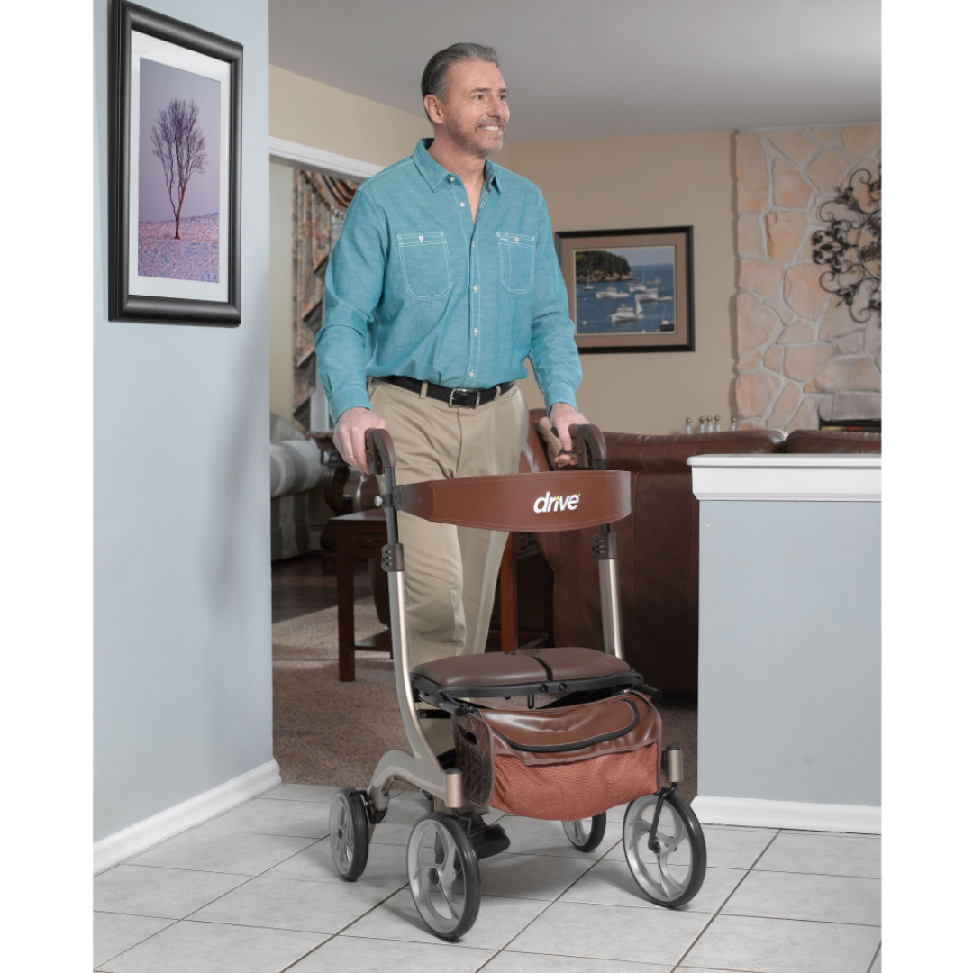 A smiling man in a teal shirt uses the Drive Nitro® DLX Rollator with seat and storage bag inside his home, confidently walking on a tiled floor with a cozy living room in the background—a practical mobility solution for daily support.