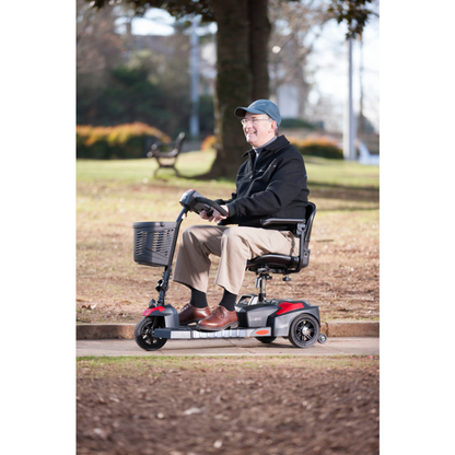 An older man in a cap and jacket smiles as he rides a Drive Scout Compact 3-Wheel Scooter along a paved park path, surrounded by trees and grass.