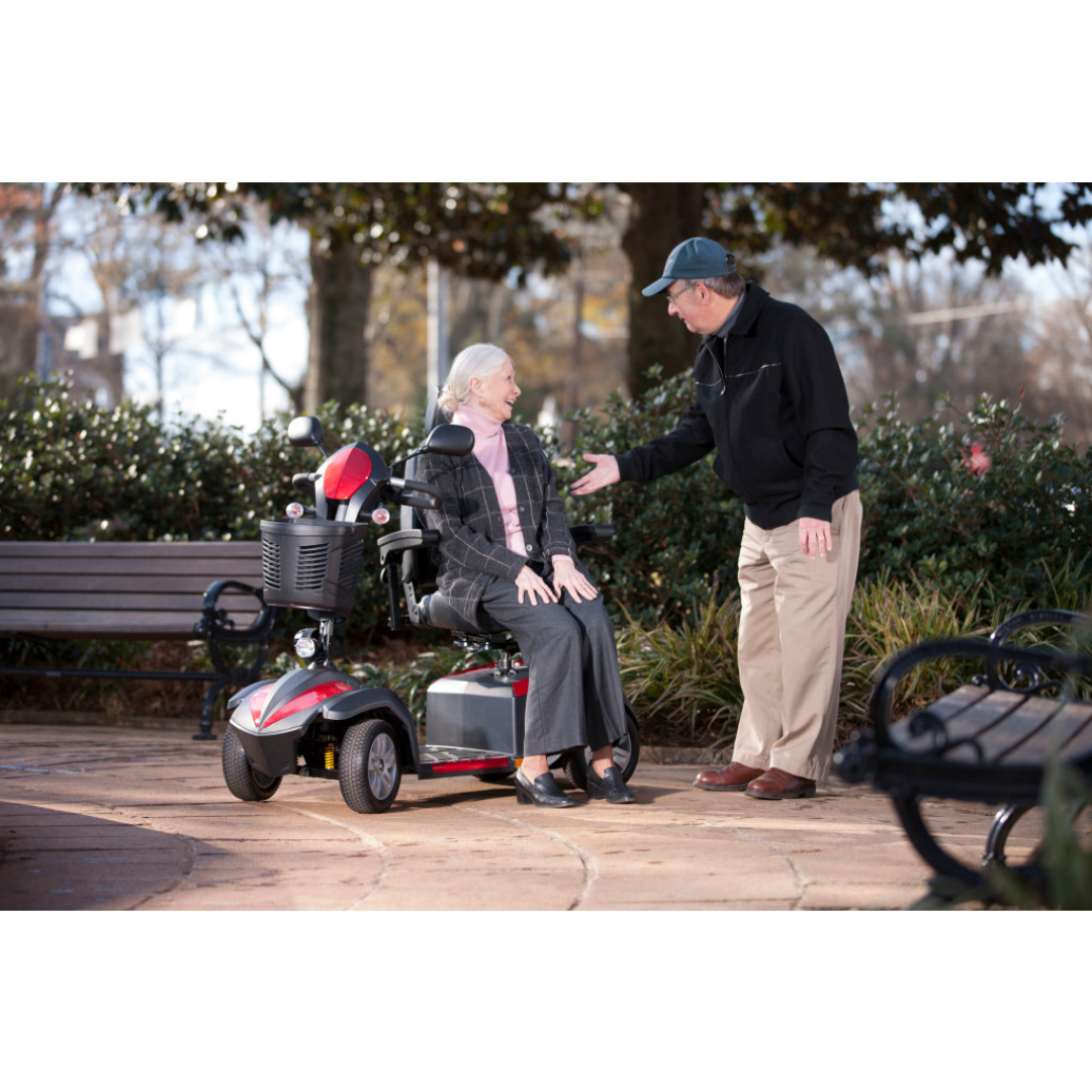 An older woman smiles while seated on a Drive Ventura 4-Wheel HD Bariatric Scooter in a park, as an older man gestures and talks to her. They are surrounded by benches, trees, and greenery.