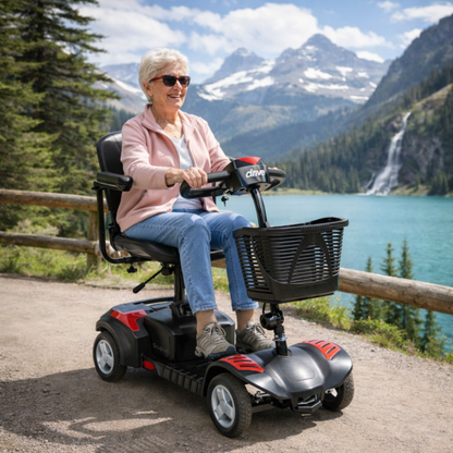 An older woman with short gray hair and sunglasses smiles as she rides a Drive Scout LT 4-Wheel Scooter along a lakeside trail, framed by snow-capped mountains, pine trees, and a waterfall in the scenic background.