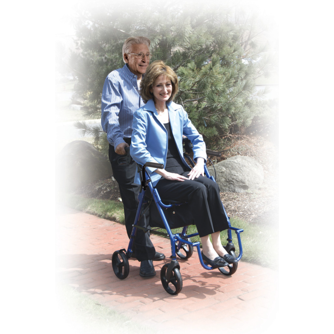 An older man smiles while pushing a woman seated in a blue Drive Hybrid Duet Rollator/Transport Chair with flip-up armrests along a brick path outdoors amid trees and rocks.