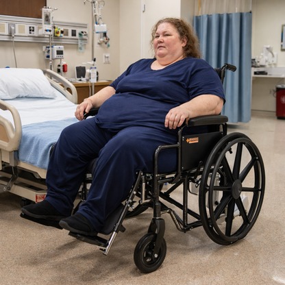A woman in a Dynarex Bariatric Wheelchair with Elevating Leg Rests sits by a hospital bed in a medical room, wearing blue. Medical equipment and Dynarex Bari+Max supplies are visible in the background.