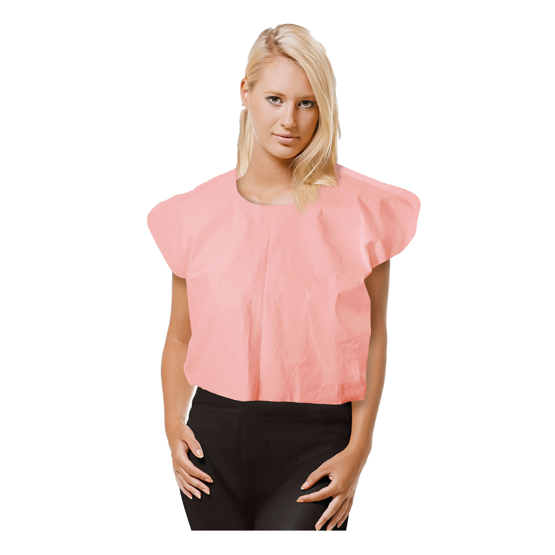 A person with blonde hair wears a Dynarex Exam Cape, a loose, short-sleeved light pink medical cape, over a black outfit while standing against a white background.
