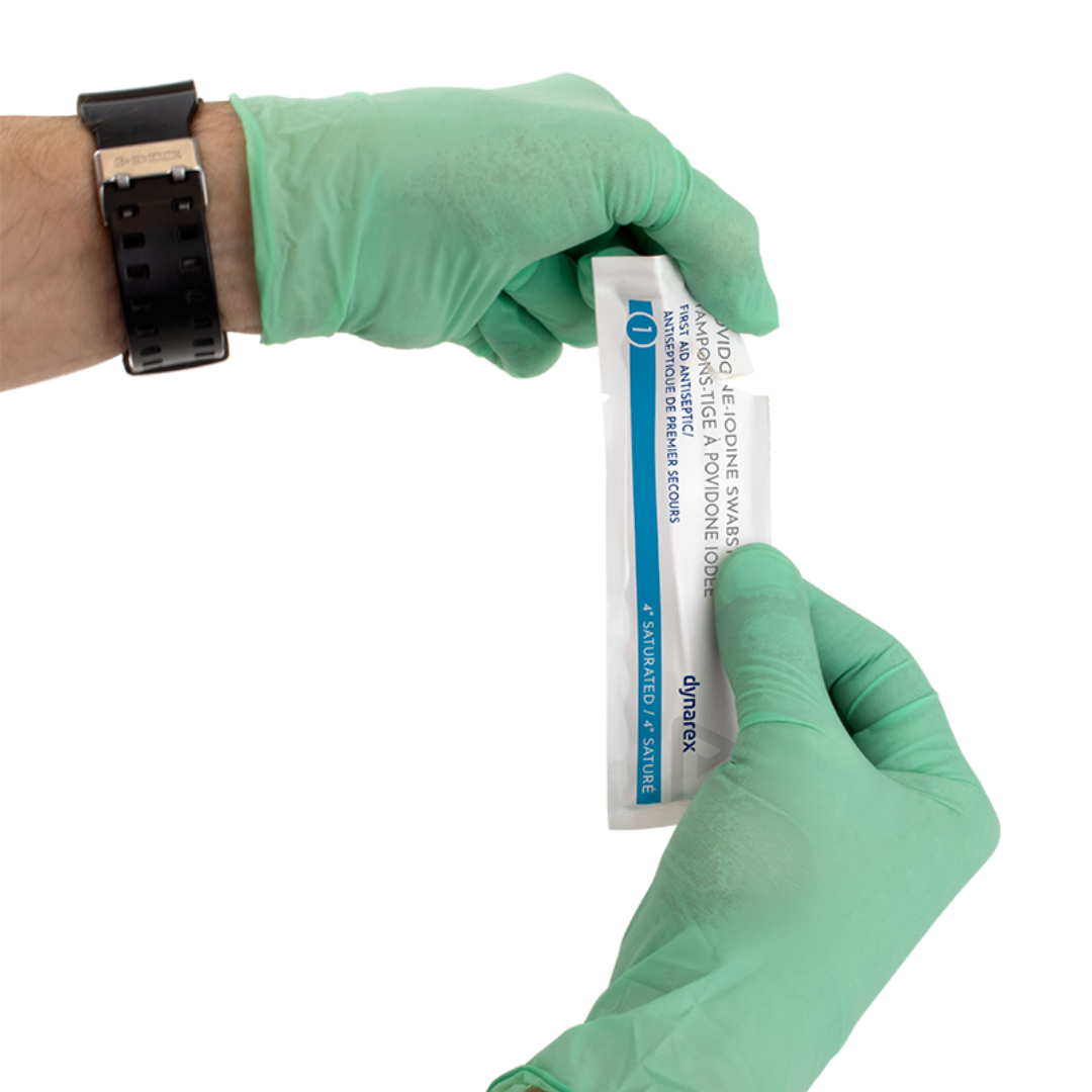 A person wearing green gloves holds an unopened Dynarex Povidone Iodine Swabsticks packet, commonly used for antiseptic skin prep, gripping each end of the packaging.