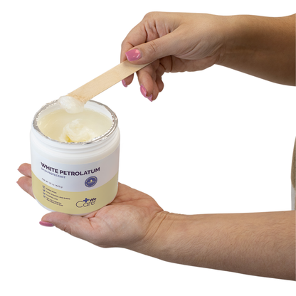 A person holds a jar of Dynarex White Petrolatum – Petroleum Jelly Skin Protectant in one hand and uses a wooden spatula with the other to scoop some out, ready to soothe dry skin.