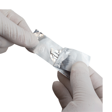 Gloved hands are opening a small metallic foil packet, revealing a thin silver Dynarex Medi-Cut Surgical Blades - Sterile against a plain white background.