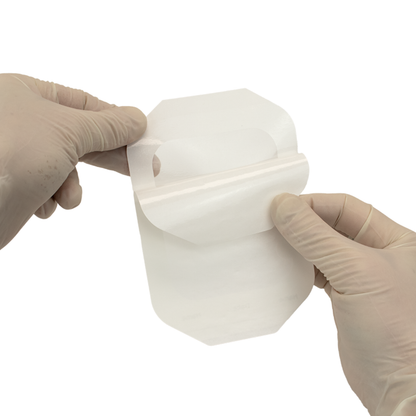 Gloved hands peel the backing off a Dynarex Transparent Film Dressing Frame Style, preparing it for use as a catheter site protector on a plain white background, highlighting its wound protection function.