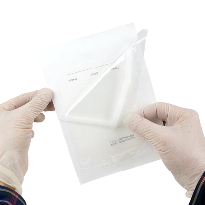 Gloved hands peel the backing off a Dynarex Transparent Film Dressing Frame Style, used for wound or catheter site protection, from its packaging against a white background.