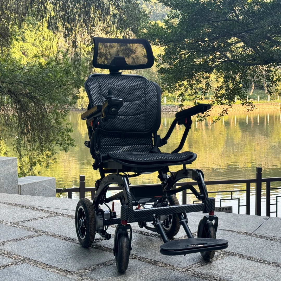 The EASWE B10 Portable Folding Powerchair - Airline Approved, a modern black electric wheelchair with dual motors and a headrest, is parked outdoors on a stone pathway near a calm lake surrounded by trees and greenery.