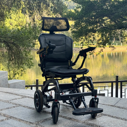 The EASWE B10 Portable Folding Powerchair - Airline Approved, a modern black electric wheelchair with dual motors and a headrest, is parked outdoors on a stone pathway near a calm lake surrounded by trees and greenery.