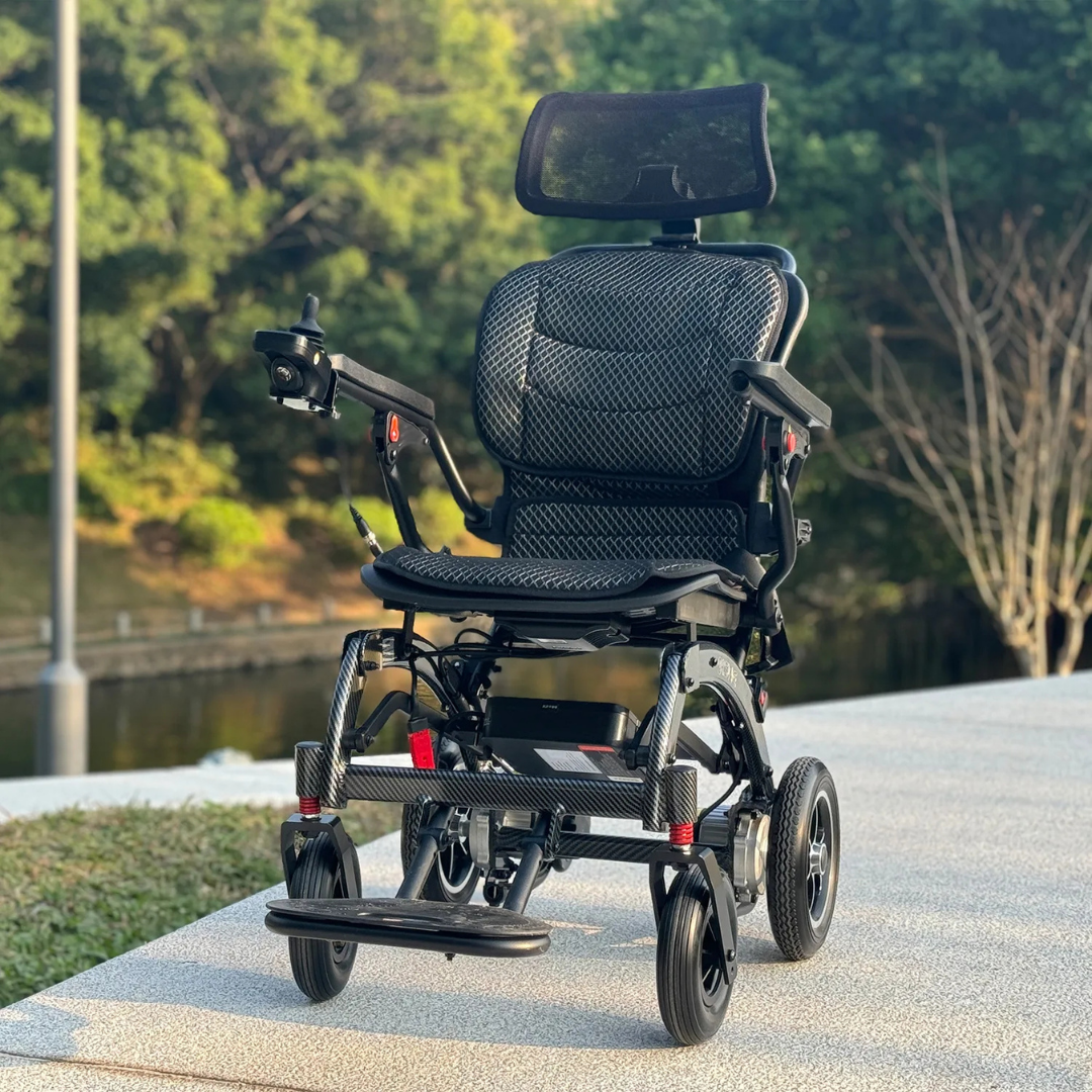 The EASWE B10 Portable Folding Powerchair – Airline Approved, featuring dual motors, mesh seat, adjustable headrest, and joystick control, is parked on a paved path outdoors among trees and greenery.