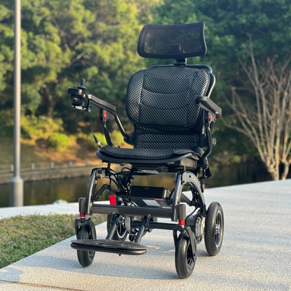 The EASWE B10 Portable Folding Powerchair – Airline Approved, featuring dual motors, mesh seat, adjustable headrest, and joystick control, is parked on a paved path outdoors among trees and greenery.