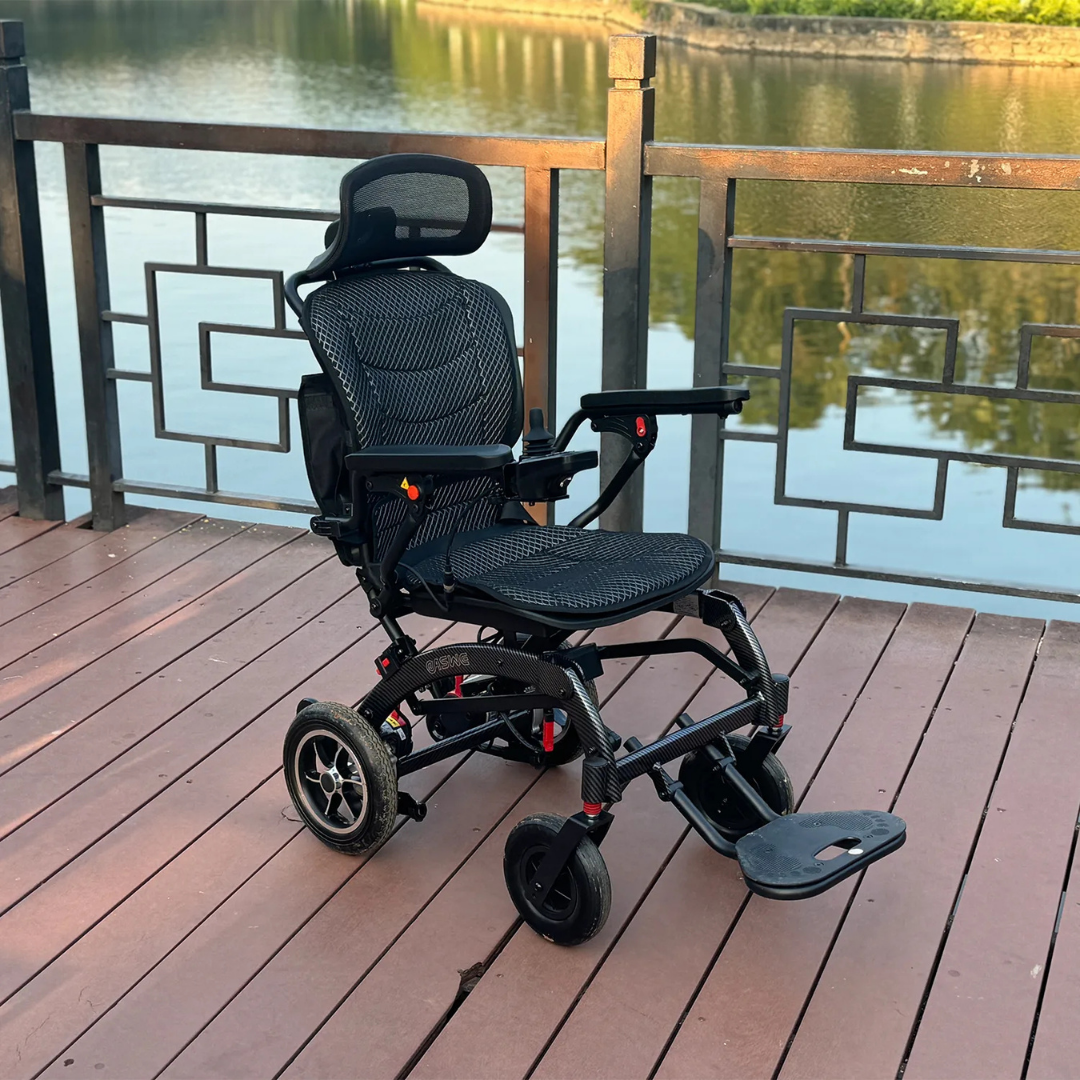 The EASWE B10 Portable Folding Powerchair - Airline Approved, in sleek black with dual motors and a mesh seat, is set on a wooden deck by a metal fence, offering views of a calm river with tree reflections.