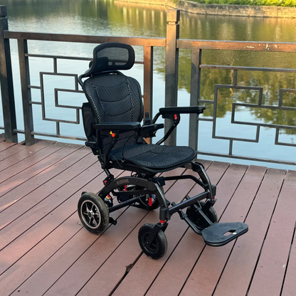 The EASWE B10 Portable Folding Powerchair - Airline Approved, in sleek black with dual motors and a mesh seat, is set on a wooden deck by a metal fence, offering views of a calm river with tree reflections.
