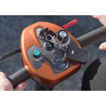 Close-up of hands operating the EV Rider Transport AF4W Automatic Folding Mobility Scooter control panel, featuring buttons, a key, a dial, and a gauge display on its textured orange console.