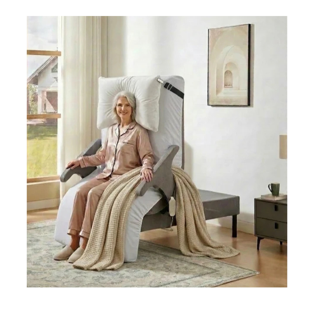 An older woman in pajamas relaxes in a reclining chair with a blanket over her legs beside an EZLift Bed™ - Deluxe Sleep To Stand Electric Bed in a cozy, light-filled living room with a rug, side table, and framed wall art.