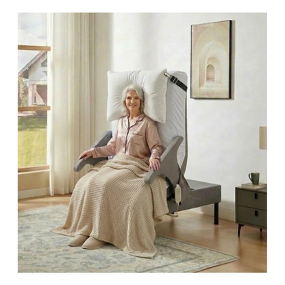 An elderly woman sits upright in the EZLift Bed™ - Deluxe Sleep To Stand Electric Bed, supported by armrests and covered in a beige blanket, smiling in a bright, neatly furnished room with a window and bedside table.