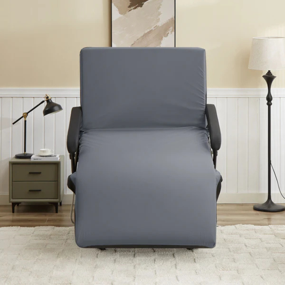 A spacious gray recliner sits in a cozy living room by a modern nightstand and lamp, next to the EZLift Bed Hi-Lo Pro™ Electric Bed with Stand Assist, with art on beige walls and a white rug completing the inviting space.