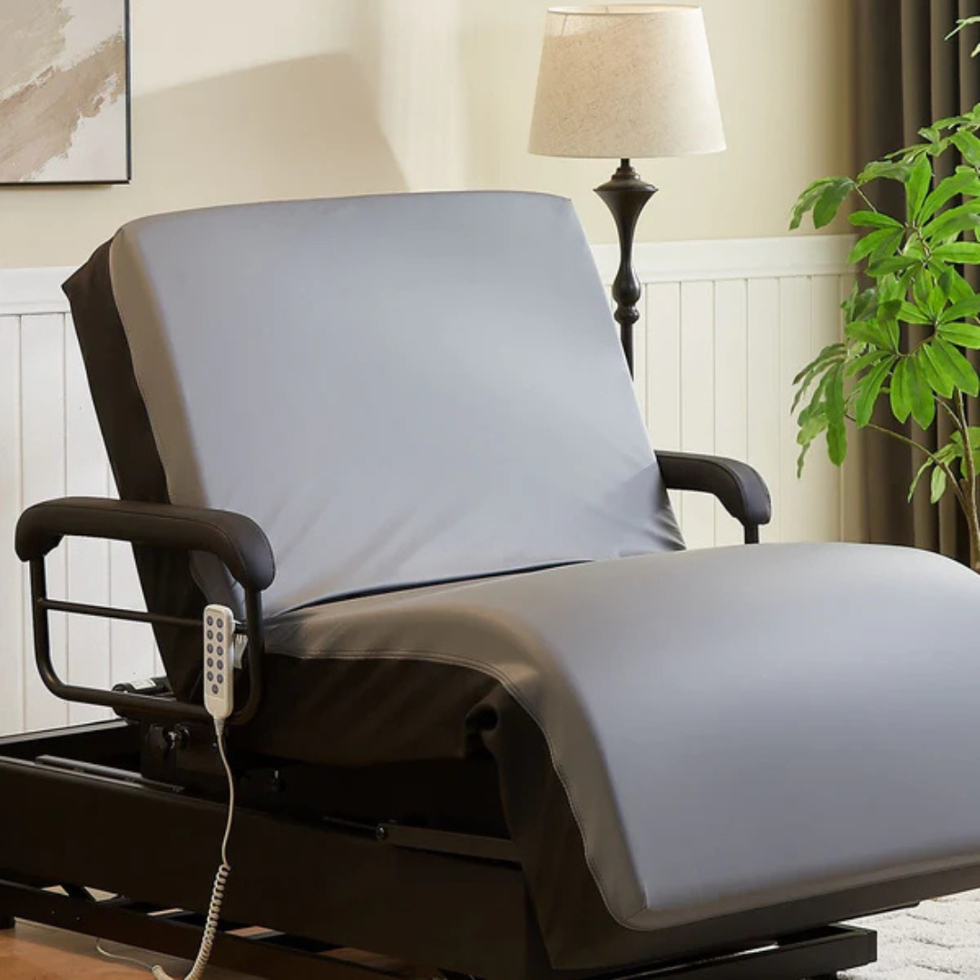 A gray adjustable medical recliner with remote sits in a cozy, well-lit room next to the EZLift Bed Hi-Lo Pro™ Electric Bed with Stand Assist, surrounded by a floor lamp, plant, and light walls with white wainscoting.