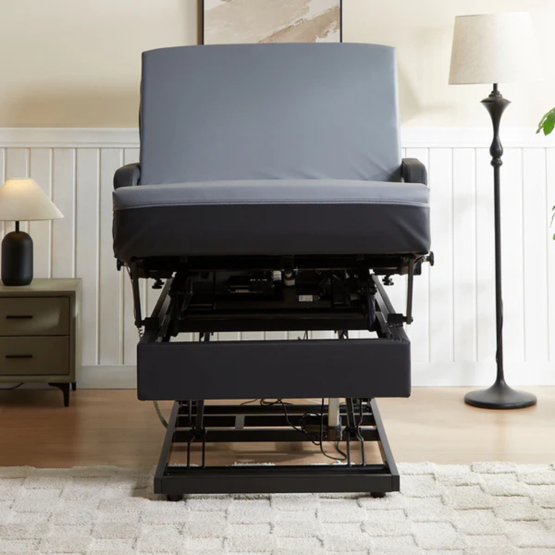 A gray upholstered reclining chair is partially elevated by a mechanical lift, similar to the EZLift Bed Hi-Lo Pro™ Electric Bed with Stand Assist. The living room has a beige rug, green nightstand with lamp, and a tall floor lamp near the wall.