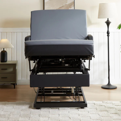 A gray upholstered reclining chair is partially elevated by a mechanical lift, similar to the EZLift Bed Hi-Lo Pro™ Electric Bed with Stand Assist. The living room has a beige rug, green nightstand with lamp, and a tall floor lamp near the wall.
