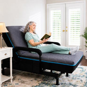 An older woman in light blue pajamas comfortably reclines on the EZLift Bed™ - Sleep To Stand Bed, reading a book as natural light fills the room. A lamp-lit nightstand beside her shows how she can regain independence with this bed.